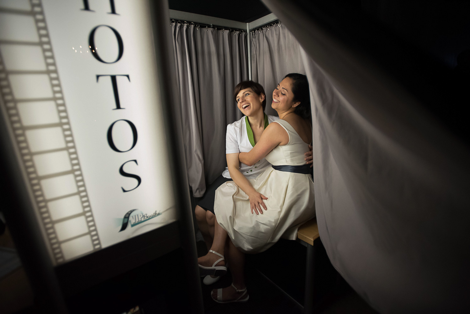 same sex couple in a sitting together, laughing and hugging in a photo booth at their wedding reception at Neu Neu