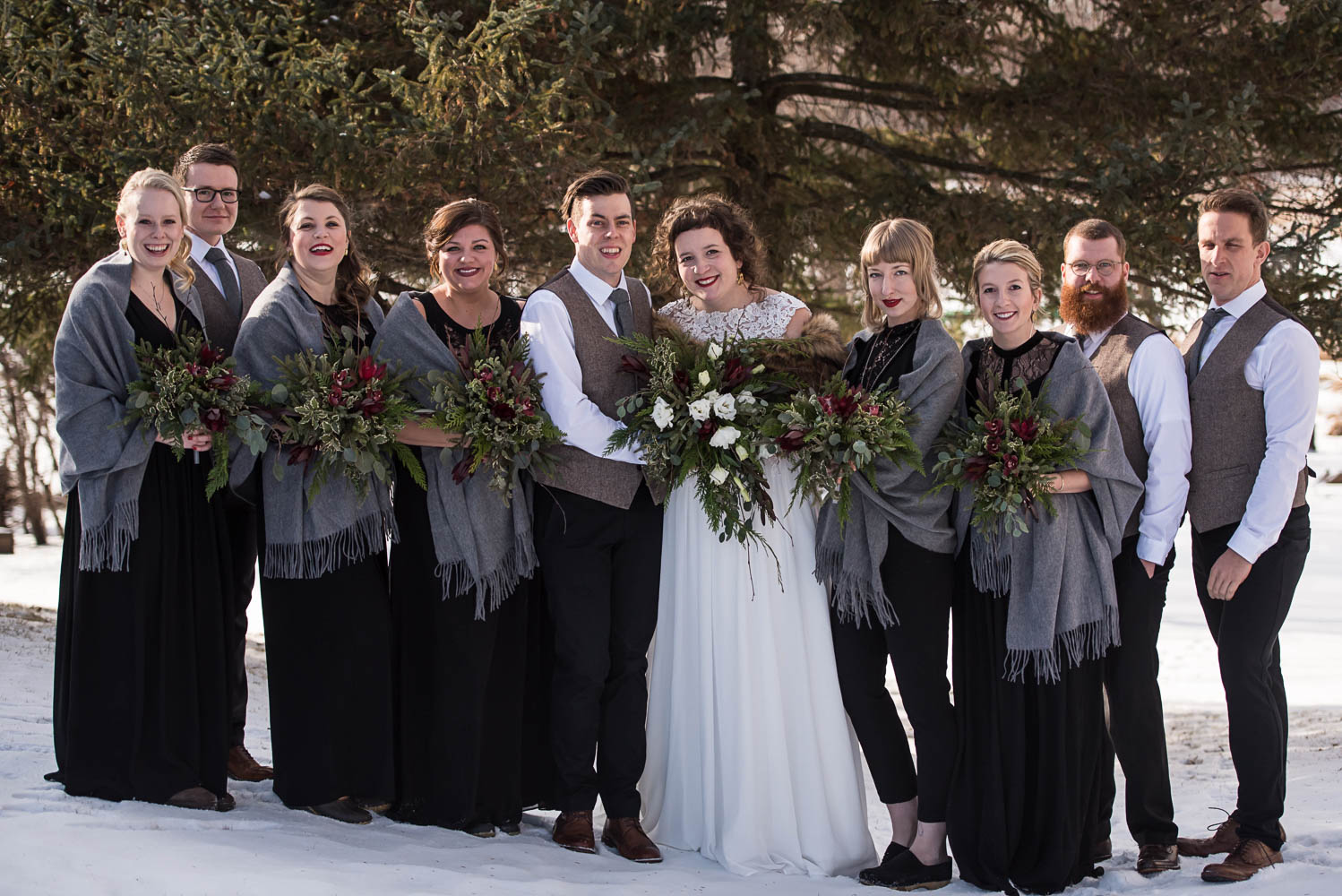 bride and groom with the bridesmaids wearing black with grey shawls holding winter greens with red and white flowers 