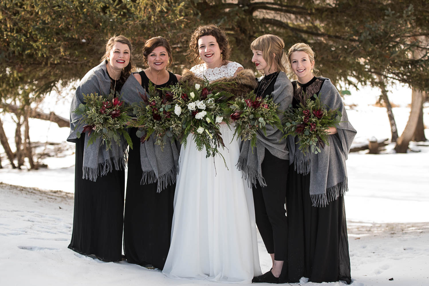 bridesmaids wearing black dresses with grey shawls standing with the bride in the snow before the winter wedding in Minnesota