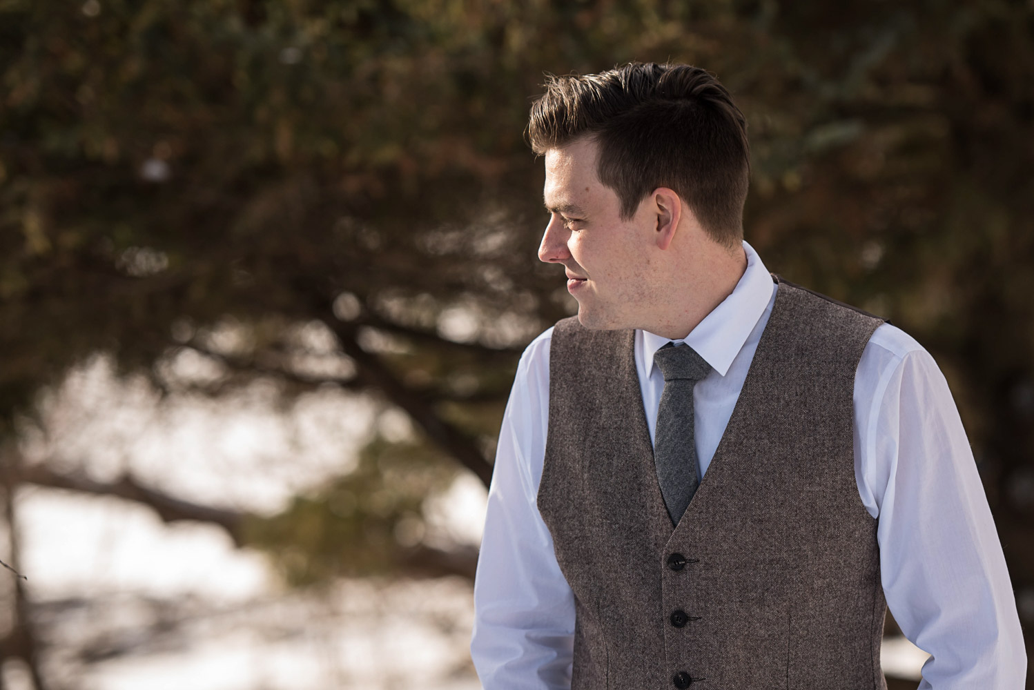 groom standing outside in the snow next to a pine tree wearing a vest and green tie