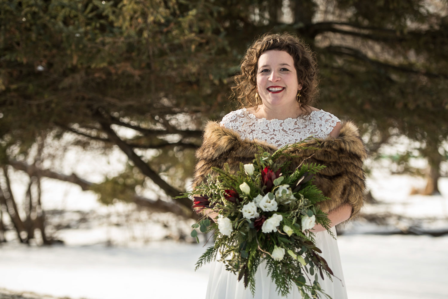 bride wearing a fur cape holding her bouquet standing next to a pine tree in the snow
