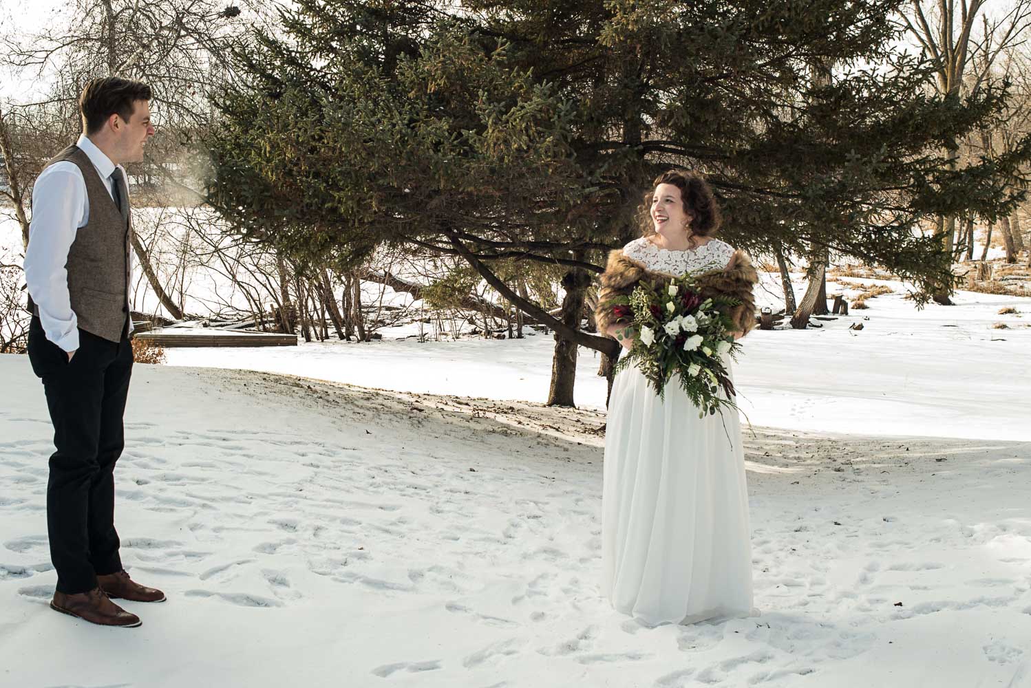 bride in a white lace dress with a fur cape holding a bouquets of white roses and evergreens before the Minnesota winter wedding