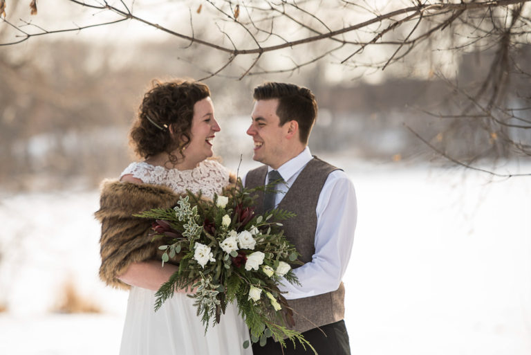 bride wearing a fur cape holding her winter bouquet laughing with the groom before their Minnesota winter wedding