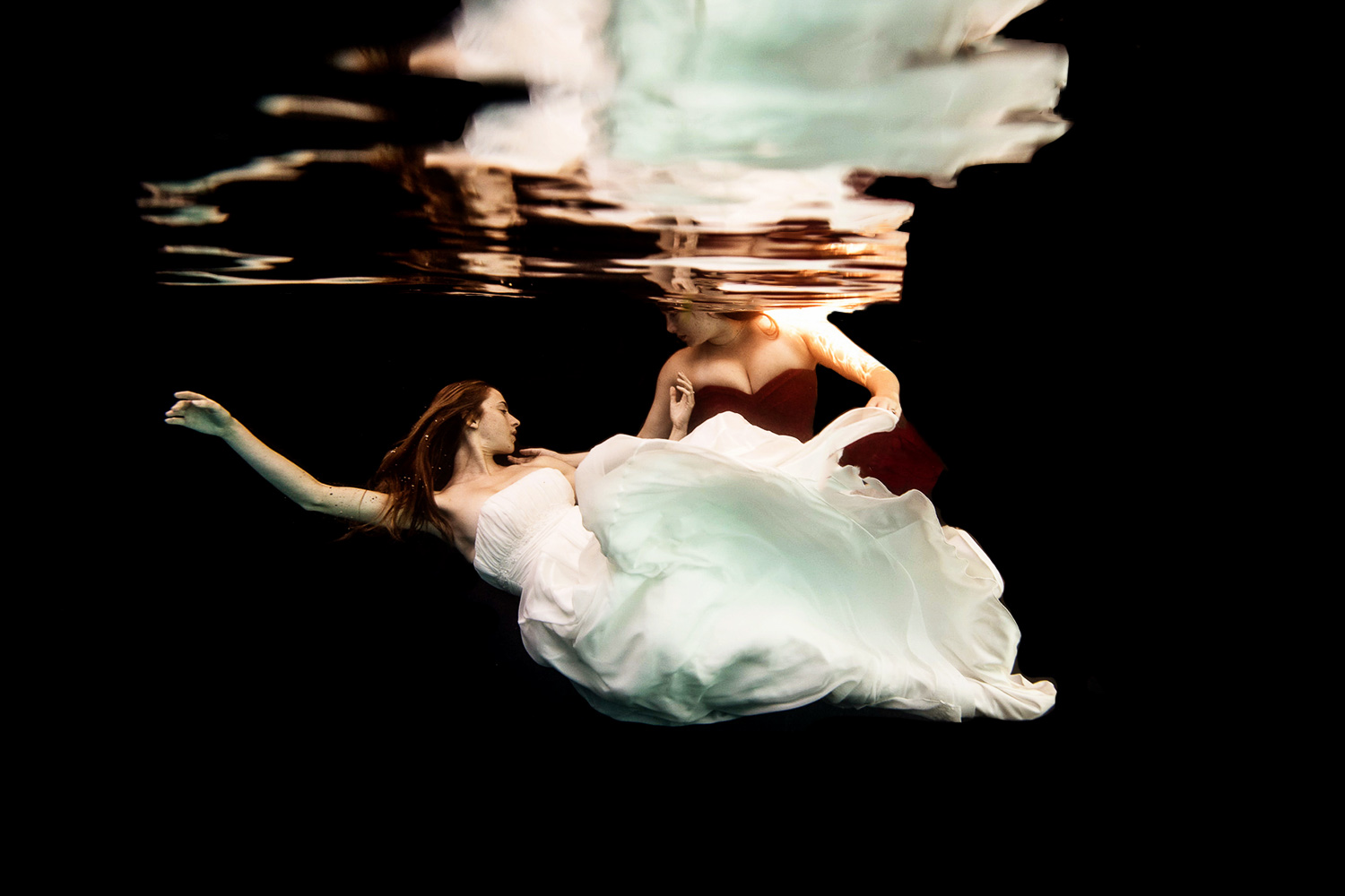 two women underwater one in a white wedding dress and the other in a red dress
