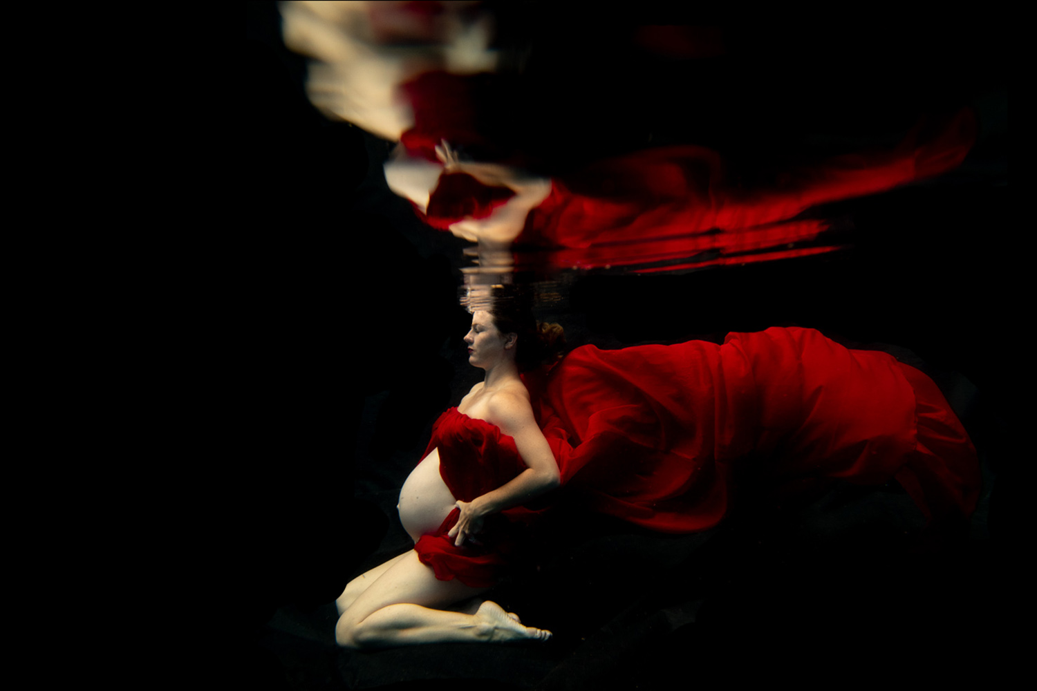 woman kneeling underwater wearing a red dress holding her belly bump