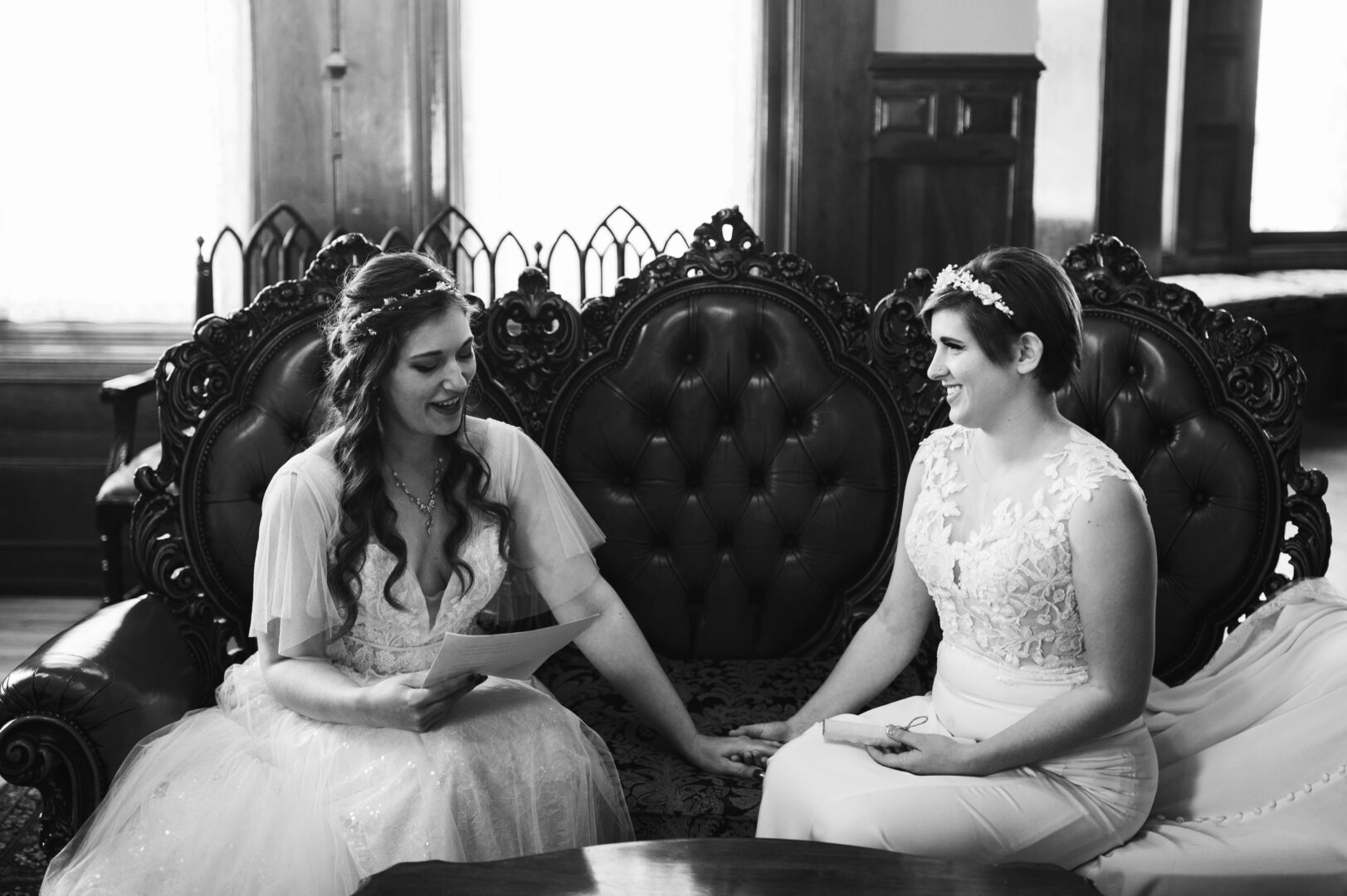 Black and white image of bride reading letter to bride while sitting on a couch and holding hands.