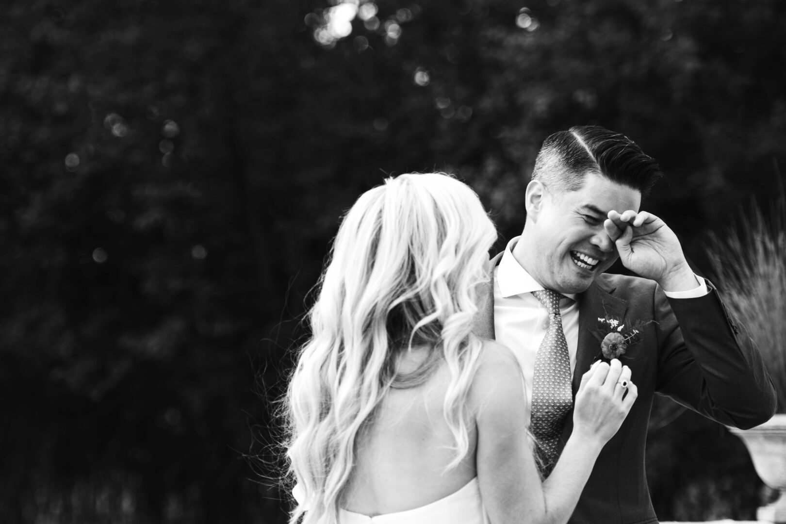 Black and white image of groom laughing and wiping his eyes during first look prior to wedding ceremony.