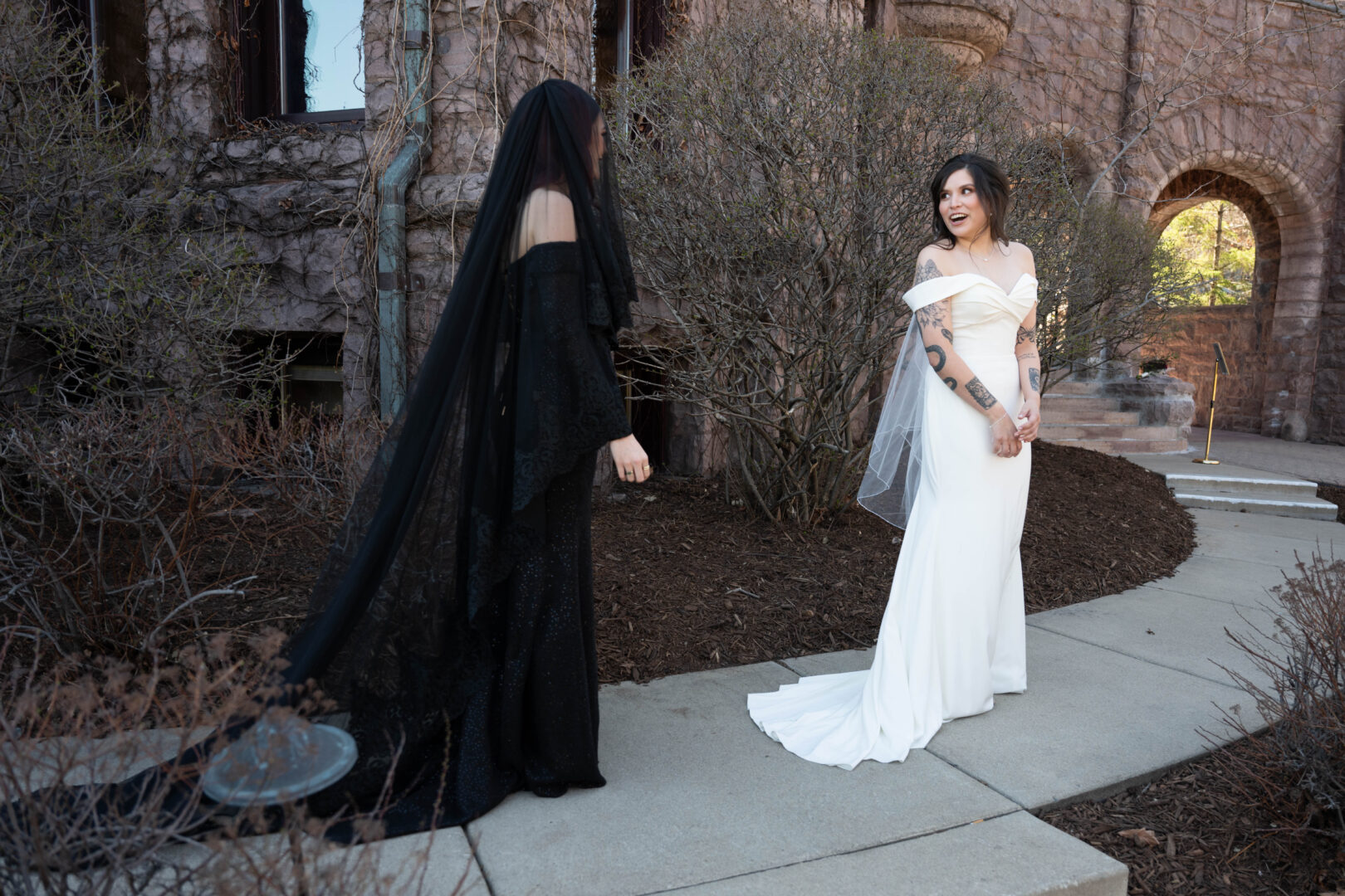 Bride in white wedding gown smiles in excitement as she sees bride in black wedding gown for the first time prior to wedding ceremony.