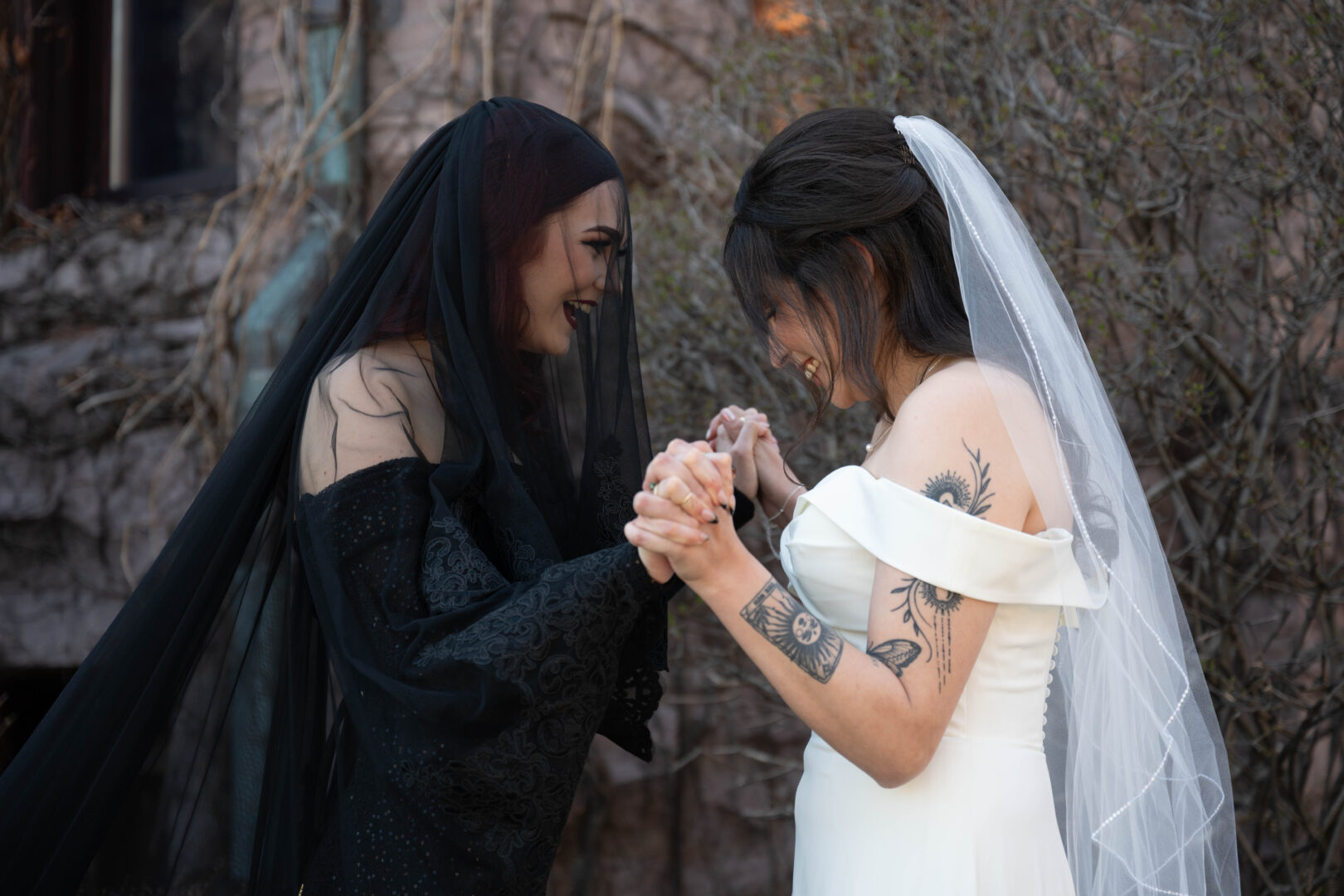 Bride in white wedding gown and bride in black wedding gown hold hands and smile at each other during first look photos prior to wedding ceremony.
